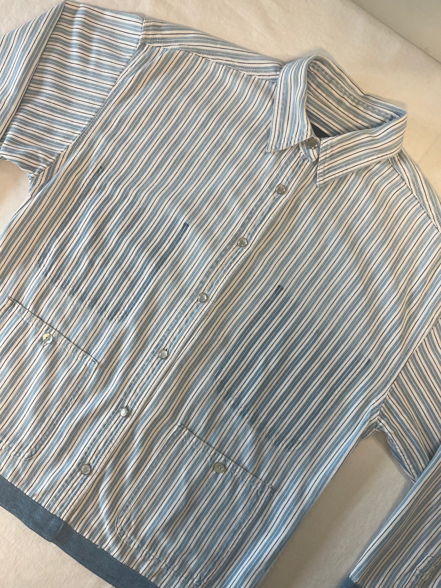 A long-sleeve button-up shirt with vertical blue, beige, and white stripes, laid flat on a light beige fabric background.